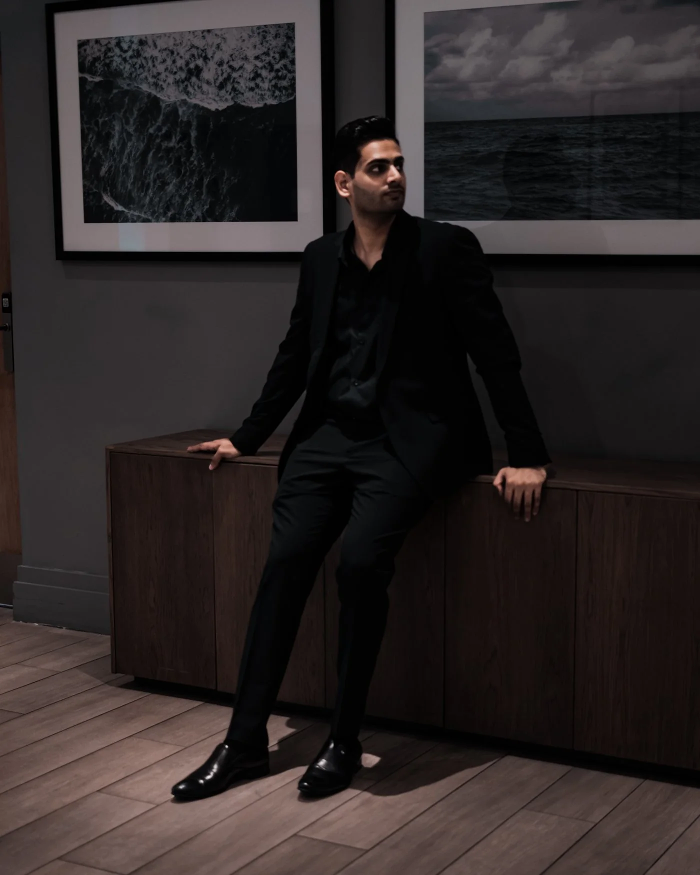 Shiv in a black suit, leaning against a wooden cabinet